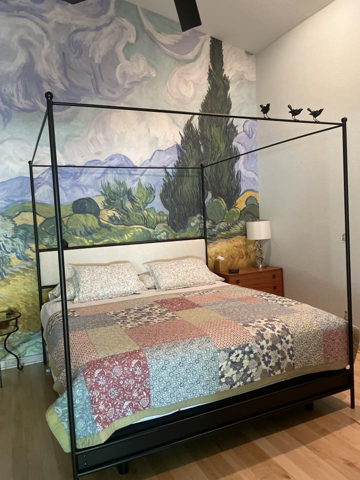 Bedroom with a bed featuring a patchwork quilt and metal canopy, against a wall with a Van Gogh-style mural.
