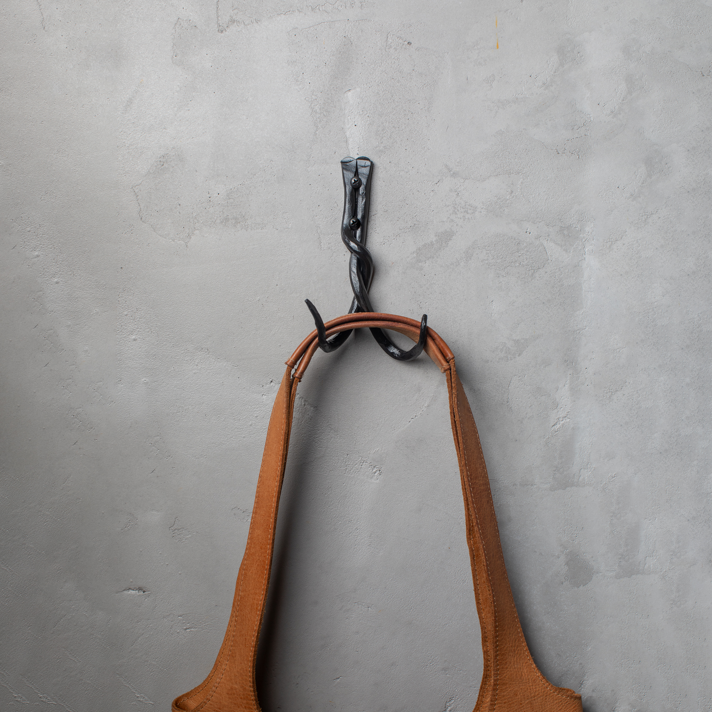 An Urban Forge Black River hand-forged iron double hook in a black finish. The hook is mounted on a gray wall and is holding a leather bag.