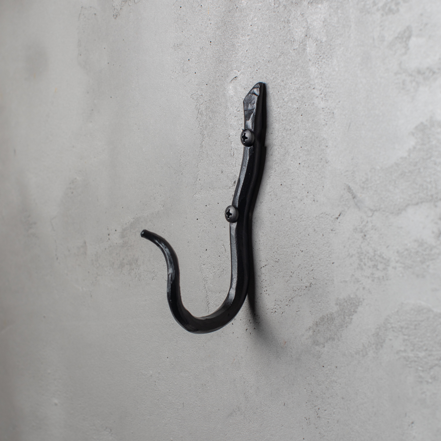 An Urban Forge Black River hand-forged iron single hook in a black finish. The hook is mounted on a gray wall.
