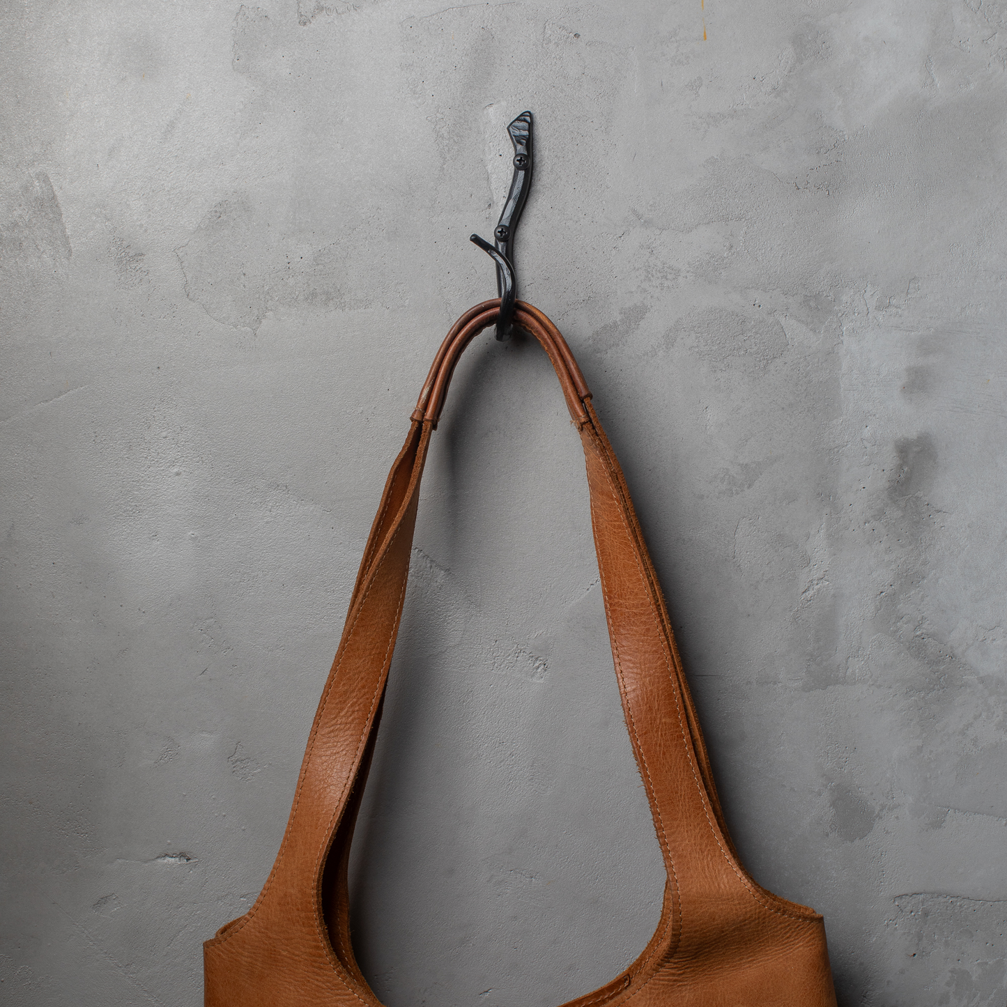 An Urban Forge Black River hand-forged iron single hook in a black finish. The hook is mounted on a gray wall and is holding a leather bag.