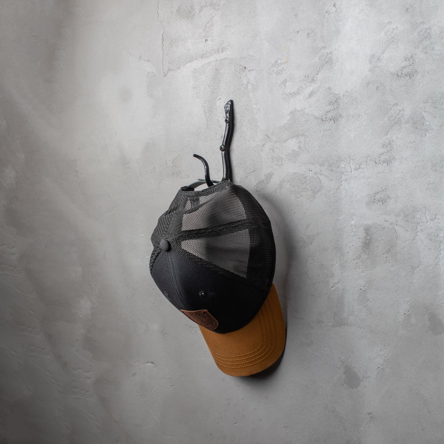 An Urban Forge Black River hand-forged iron single hook in a black finish. The hook is mounted on a gray wall and is holding a ball cap.