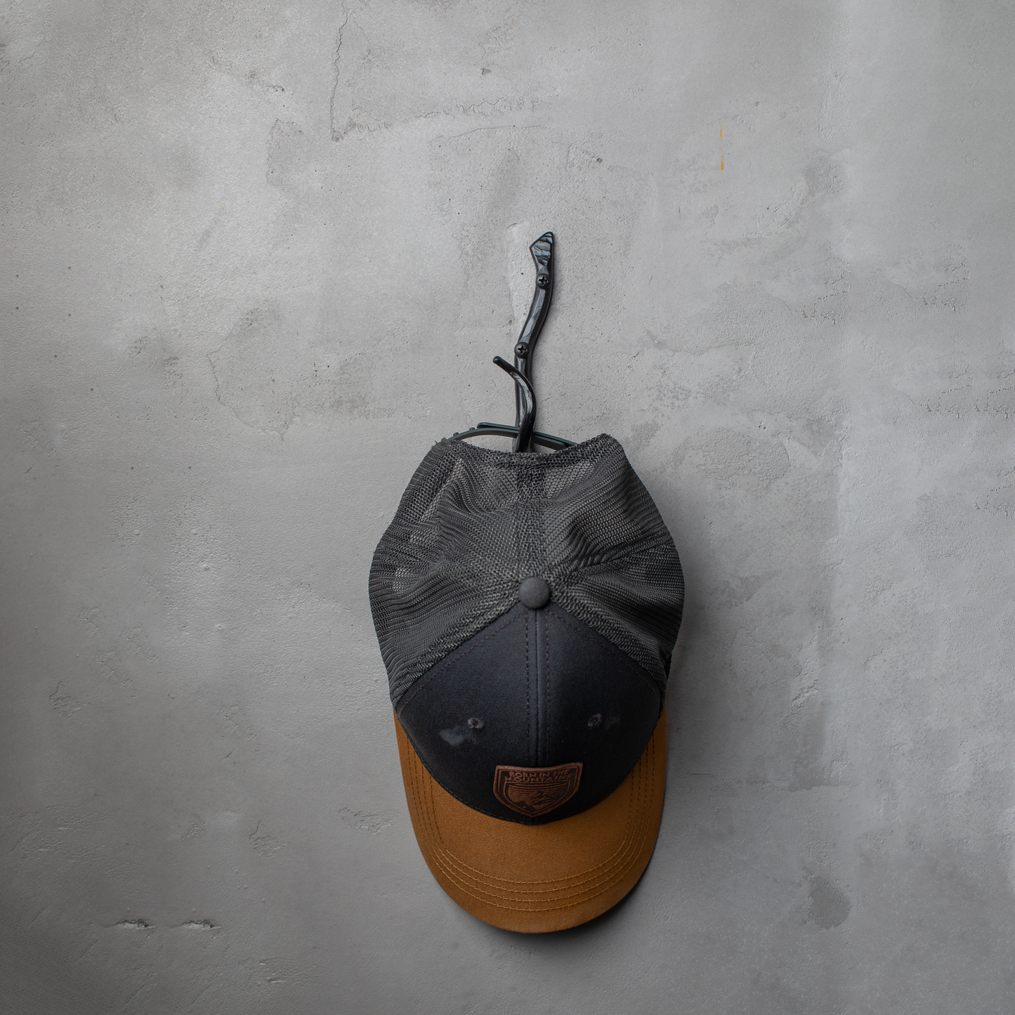 An Urban Forge Black River hand-forged iron single hook in a black finish. The hook is mounted on a gray wall and is holding a ball cap.