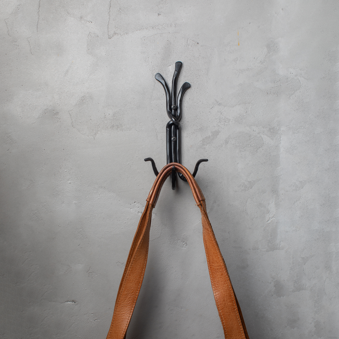 An Urban Forge Black River hand-forged iron triple hook in a black finish. The hook is mounted on a gray wall and is holding a leather bag.
