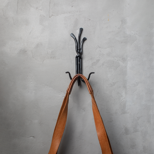 An Urban Forge Black River hand-forged iron triple hook in a black finish. The hook is mounted on a gray wall and is holding a leather bag.