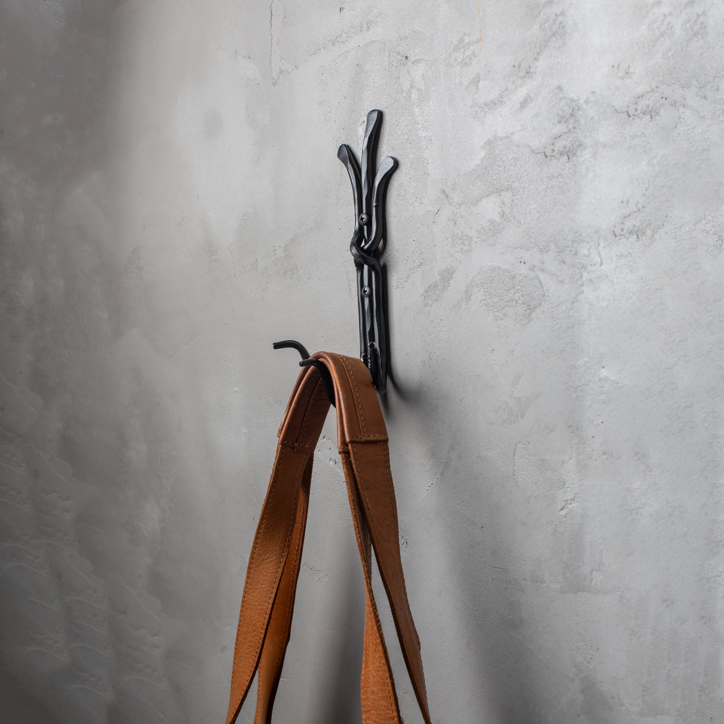 An Urban Forge Black River hand-forged iron triple hook in a black finish. The hook is mounted on a gray wall and is holding a leather bag.