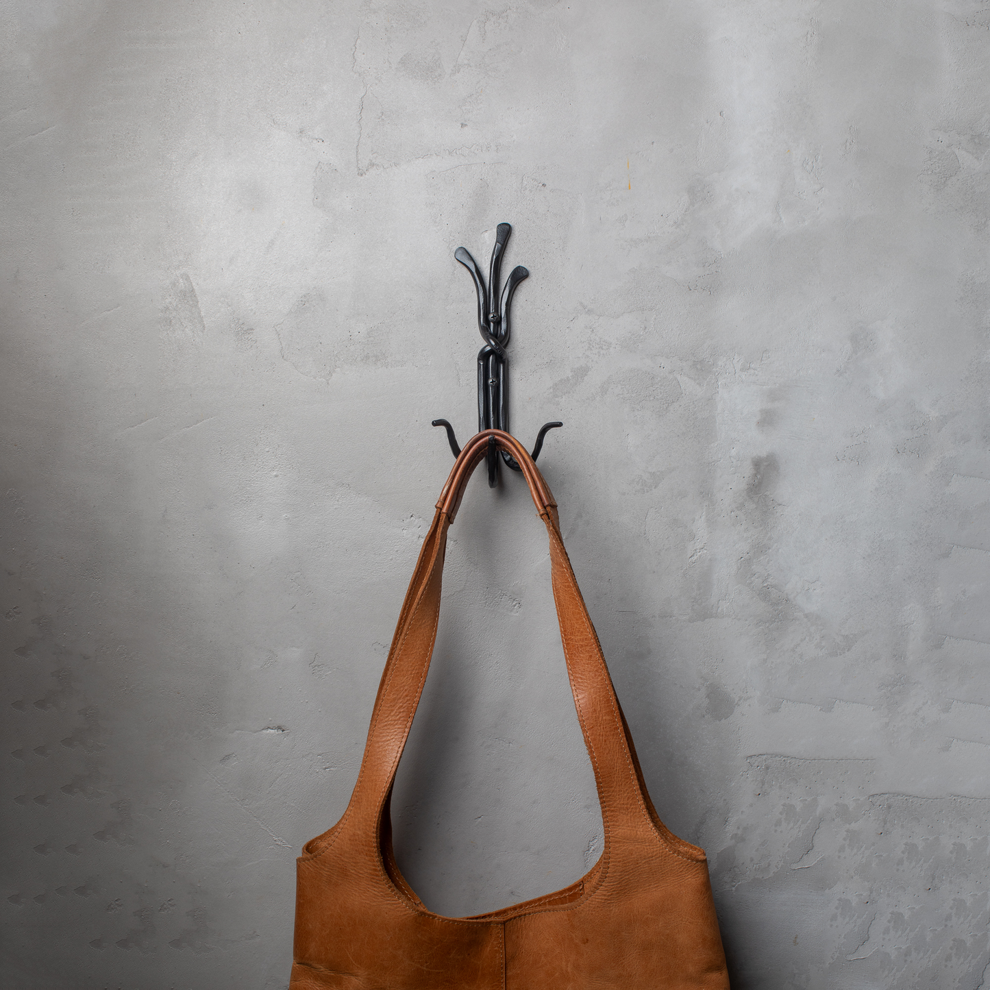 An Urban Forge Black River hand-forged iron triple hook in a black finish. The hook is mounted on a gray wall and is holding a leather bag.