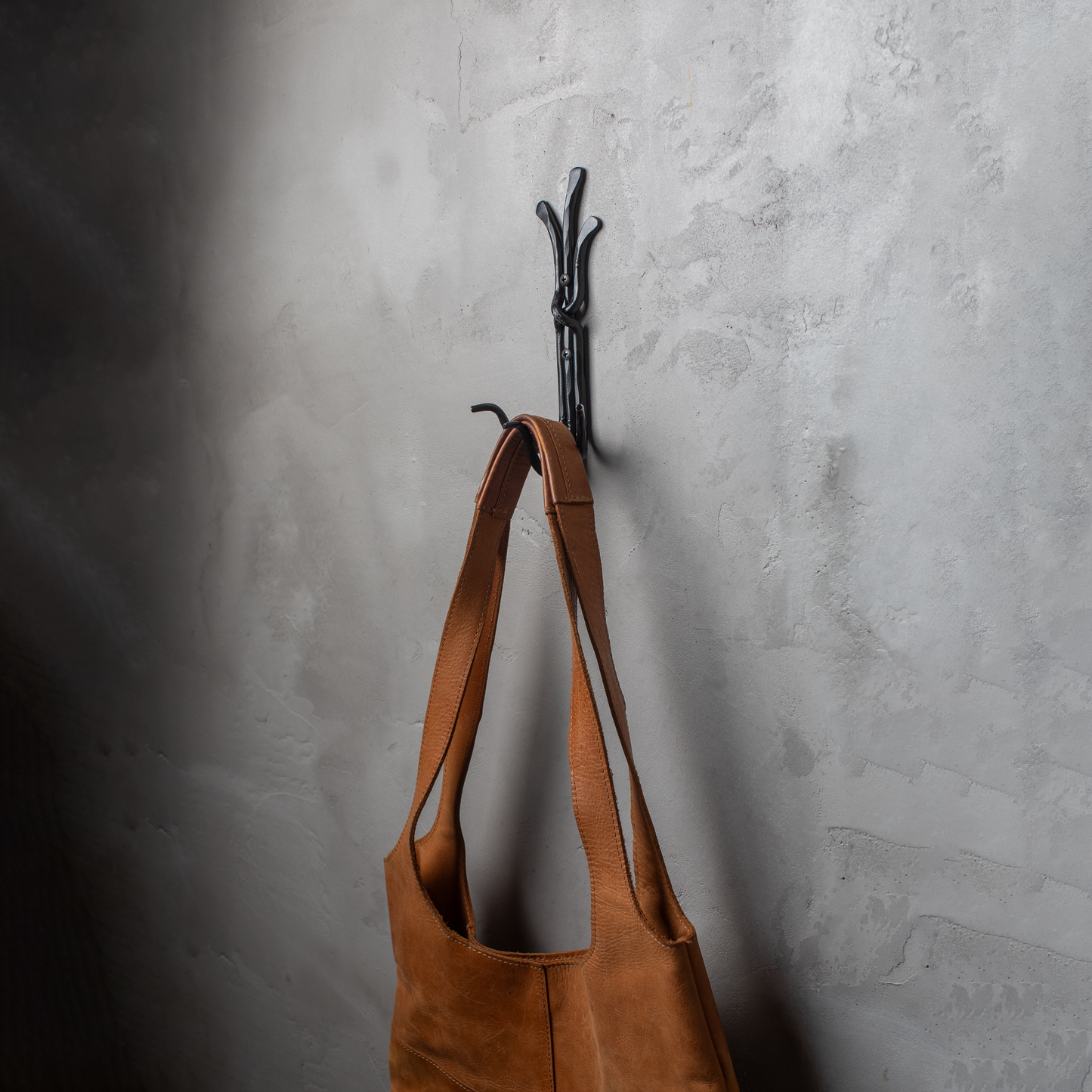 An Urban Forge Black River hand-forged iron triple hook in a black finish. The hook is mounted on a gray wall and is holding a leather bag.