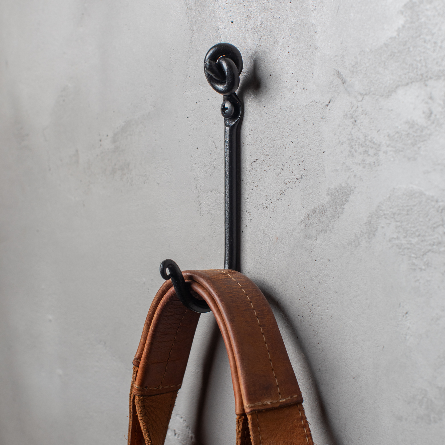 An Urban Forge Knot hand-forged iron single hook in a black finish. The hook is mounted on a gray wall and is holding a leather strap.