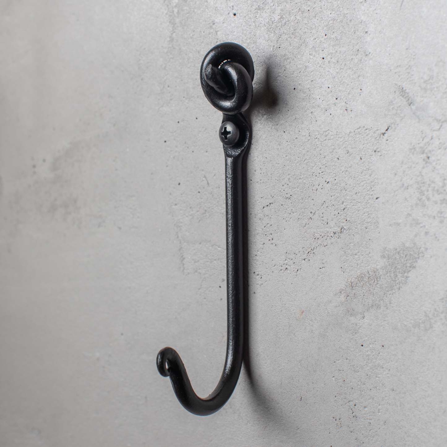 An Urban Forge Knot hand-forged iron single hook in a black finish. The hook is mounted on a gray wall.