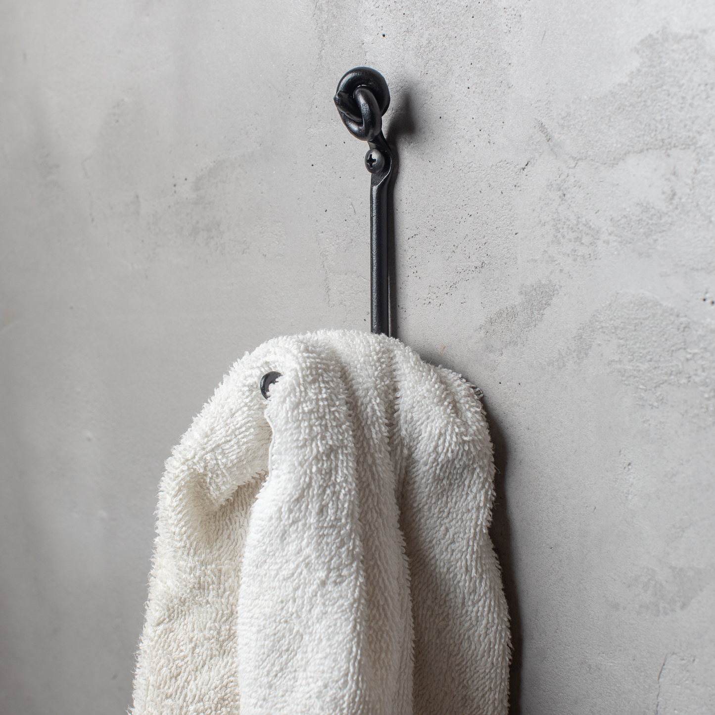 An Urban Forge Knot hand-forged iron single hook in a black finish. The hook is mounted on a gray wall and is holding a white towel.