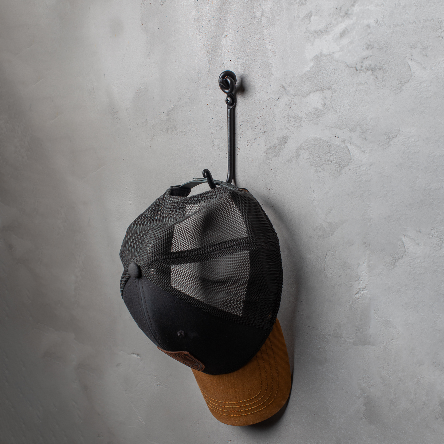 An Urban Forge Knot hand-forged iron single hook in a black finish. The hook is mounted on a gray wall and is holding a ball cap.