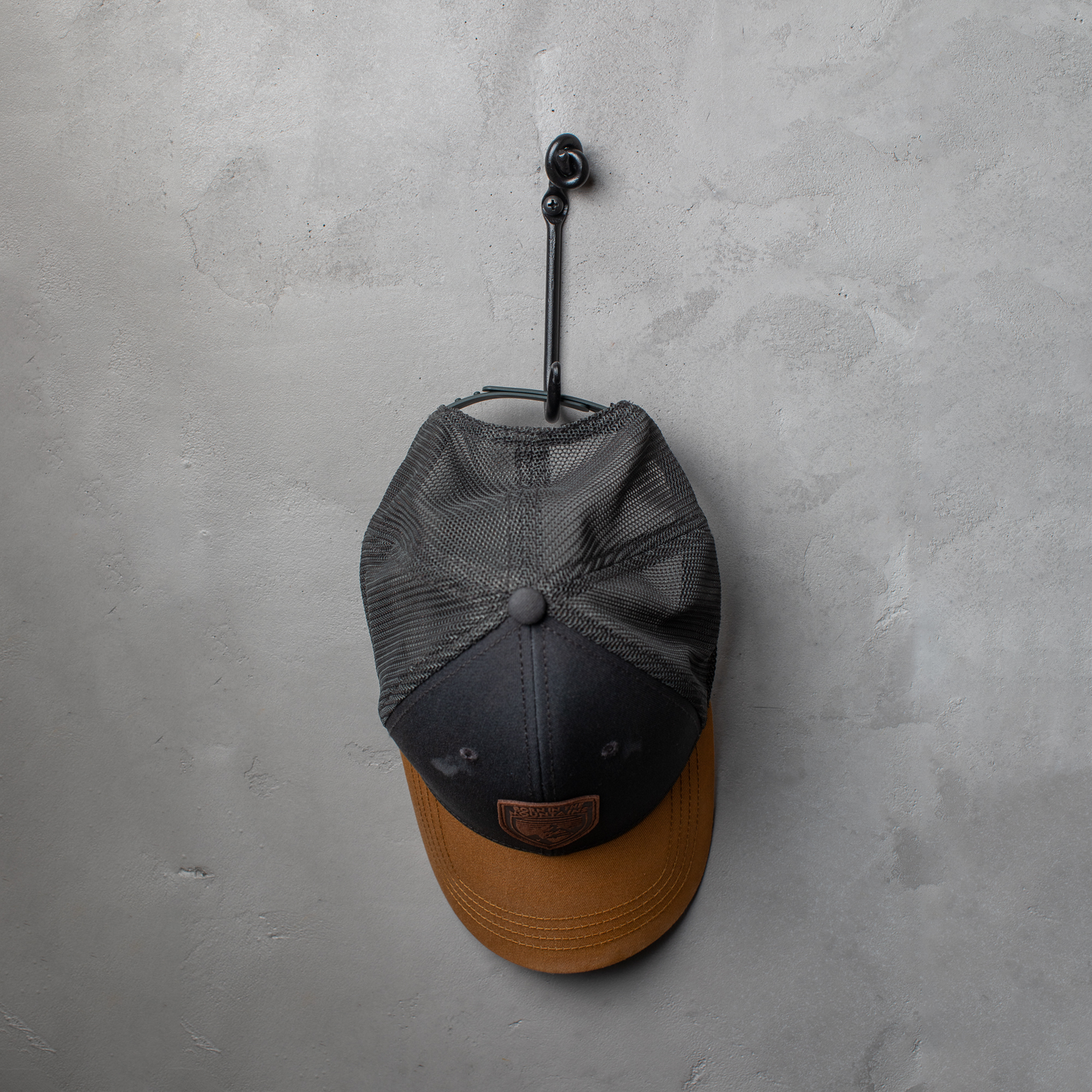 An Urban Forge Knot hand-forged iron single hook in a black finish. The hook is mounted on a gray wall and is holding a ball cap.