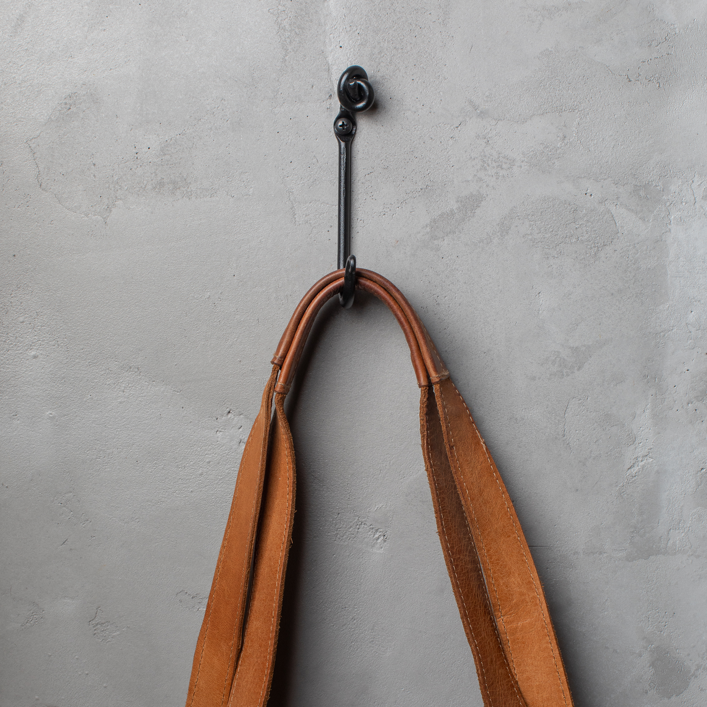 An Urban Forge Knot hand-forged iron single hook in a black finish. The hook is mounted on a gray wall and is holding a leather strap.