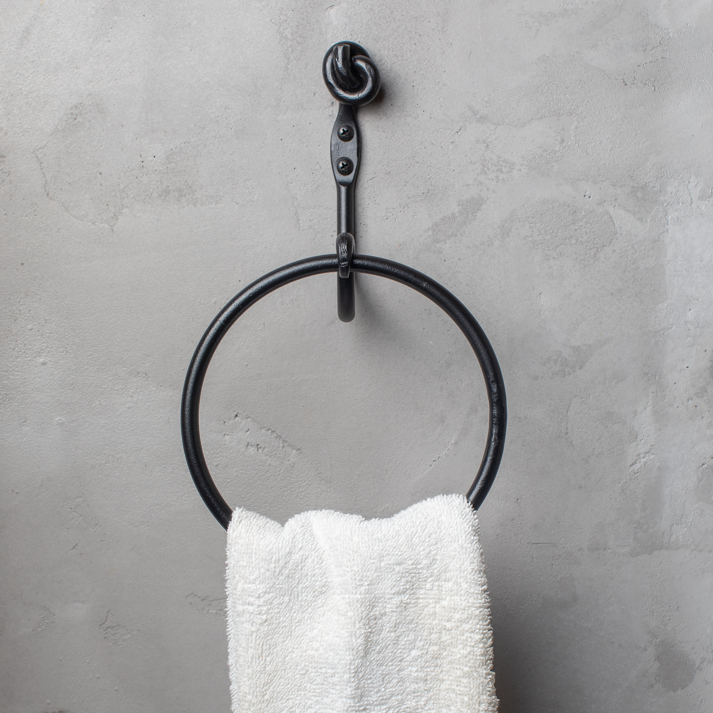 The Urban Forge Knot towel ring in a black finish. The towel ring is mounted on a gray wall and is holding a towel.
