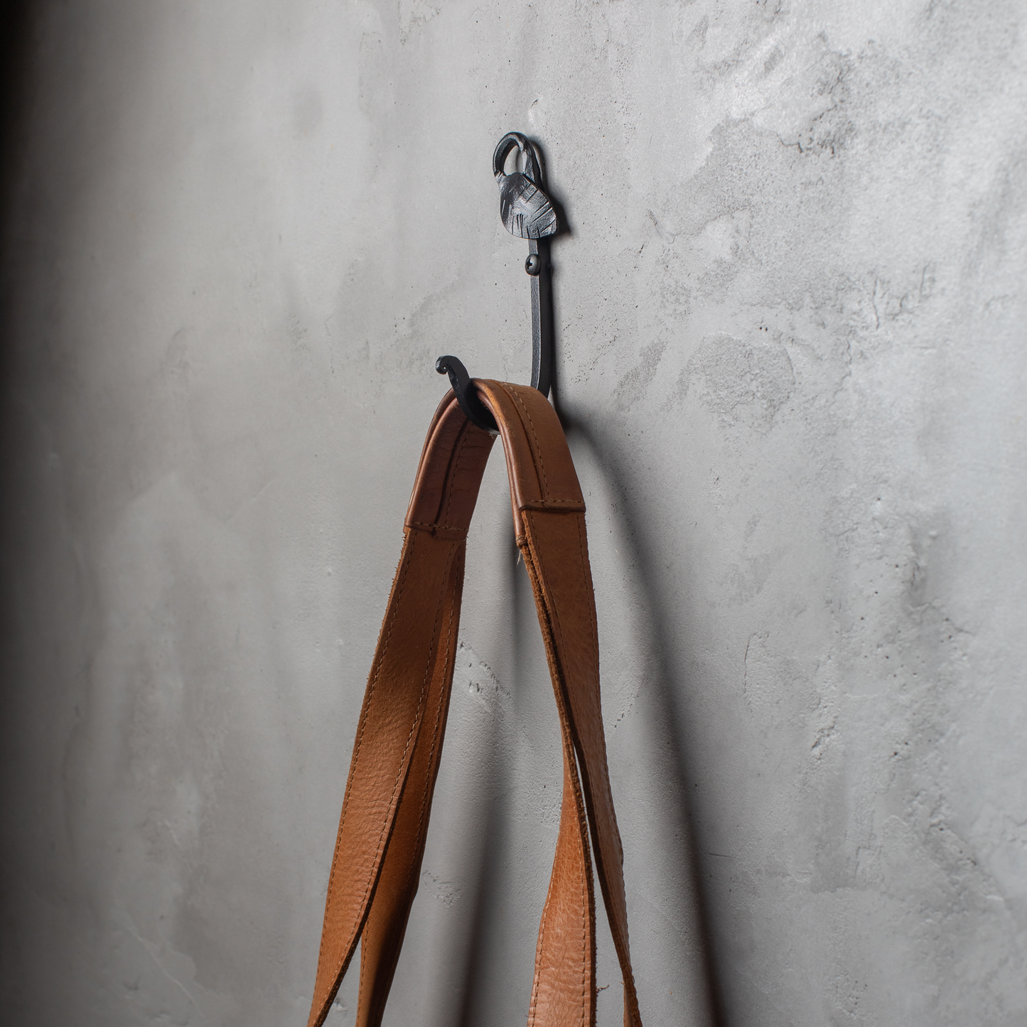 An Urban Forge Leaf hand-forged iron single hook in a black finish. The hook is mounted on a gray wall and is holding a leather bag.