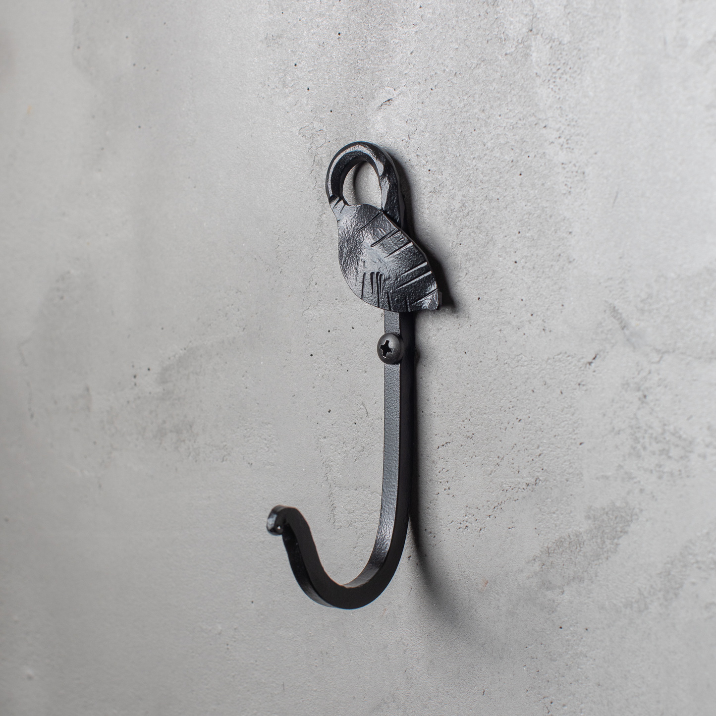 An Urban Forge Leaf hand-forged iron single hook in a black finish. The hook is mounted on a gray wall.