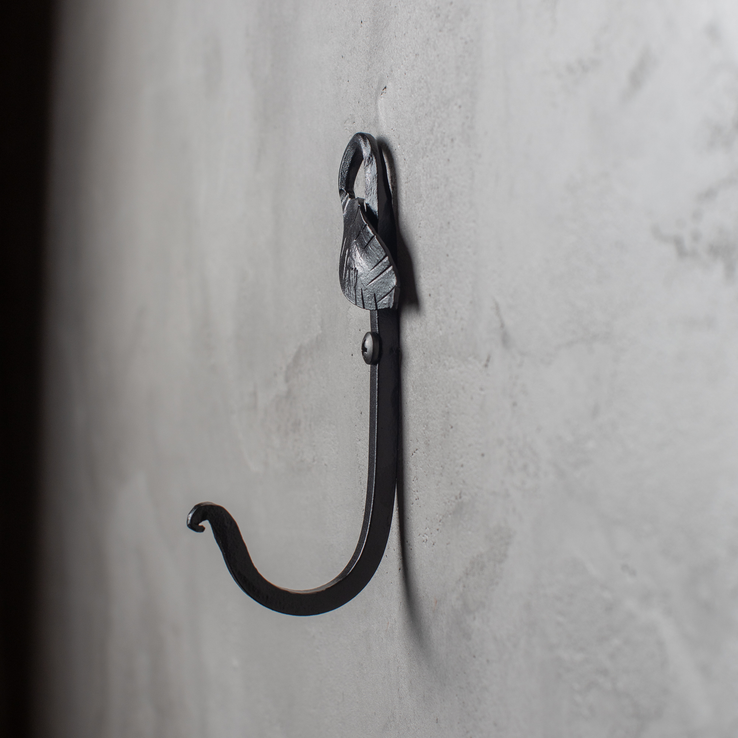 An Urban Forge Leaf hand-forged iron single hook in a black finish. The hook is mounted on a gray wall.