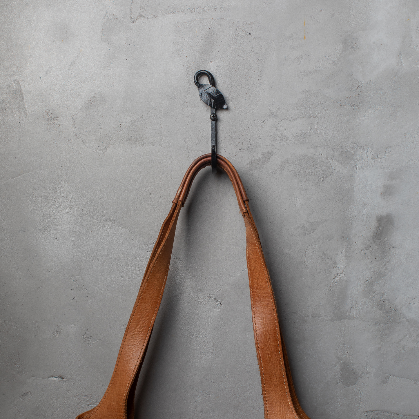 An Urban Forge Leaf hand-forged iron single hook in a black finish. The hook is mounted on a gray wall and is holding a leather bag.