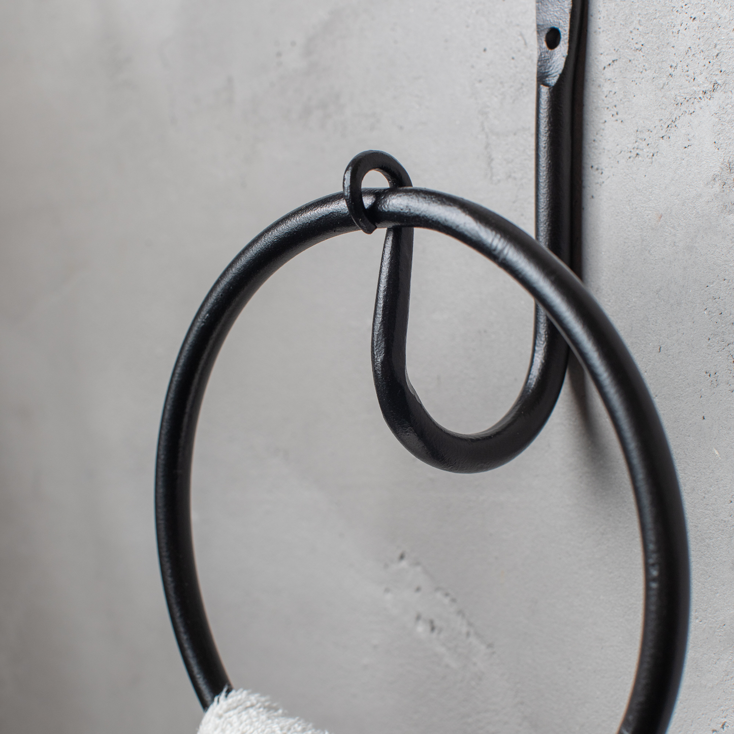 The Urban Forge Leaf towel ring in a black finish. The towel ring is mounted on a gray wall and is holding a towel.