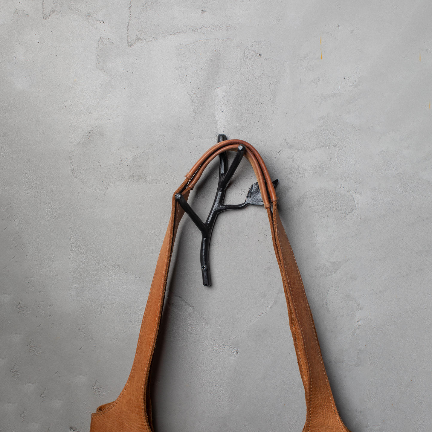 An Urban Forge Sylamore hand-forged iron double hook in a black finish. The hook is mounted on a gray wall and is holding a leather bag.