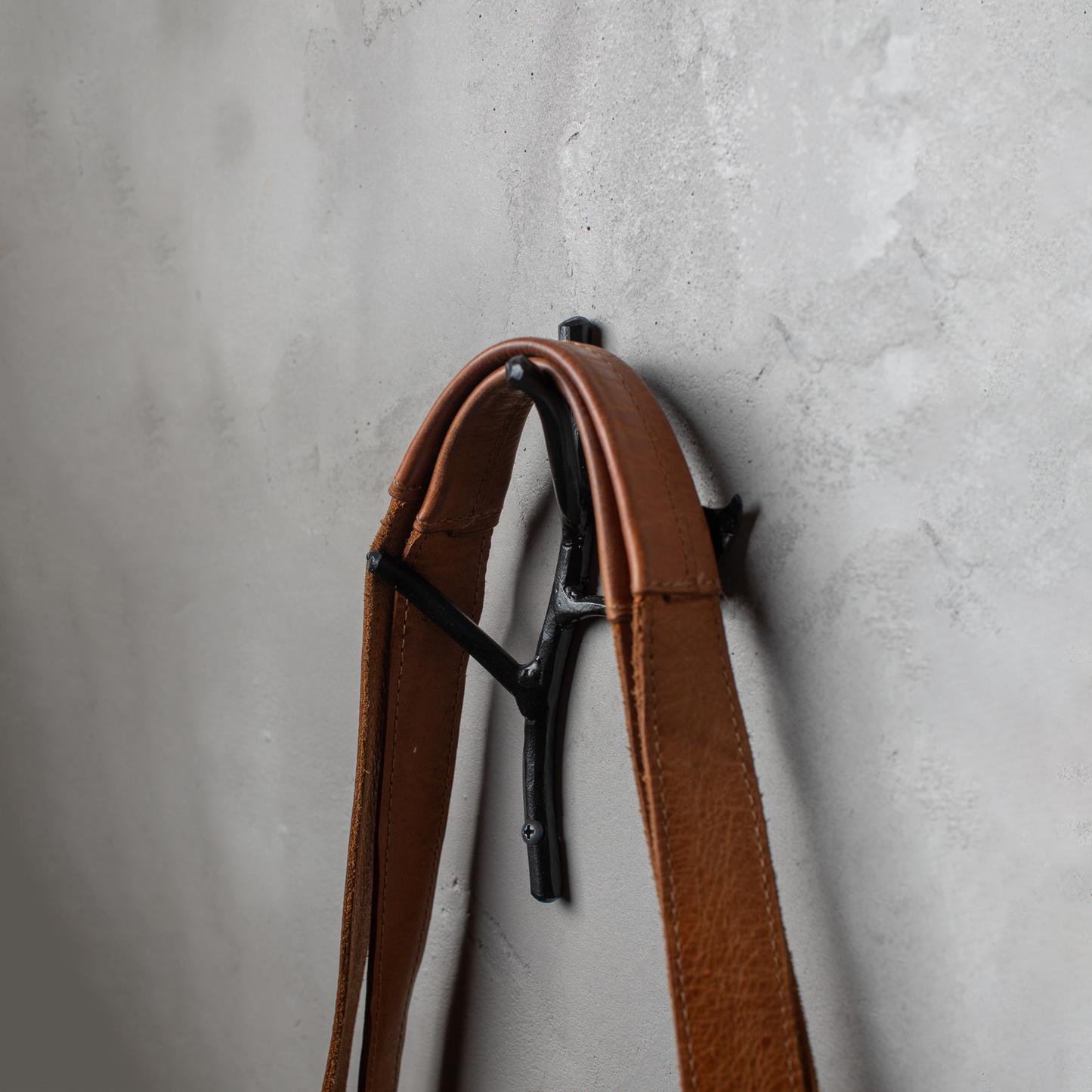 An Urban Forge Sylamore hand-forged iron double hook in a black finish. The hook is mounted on a gray wall and is holding a leather bag.
