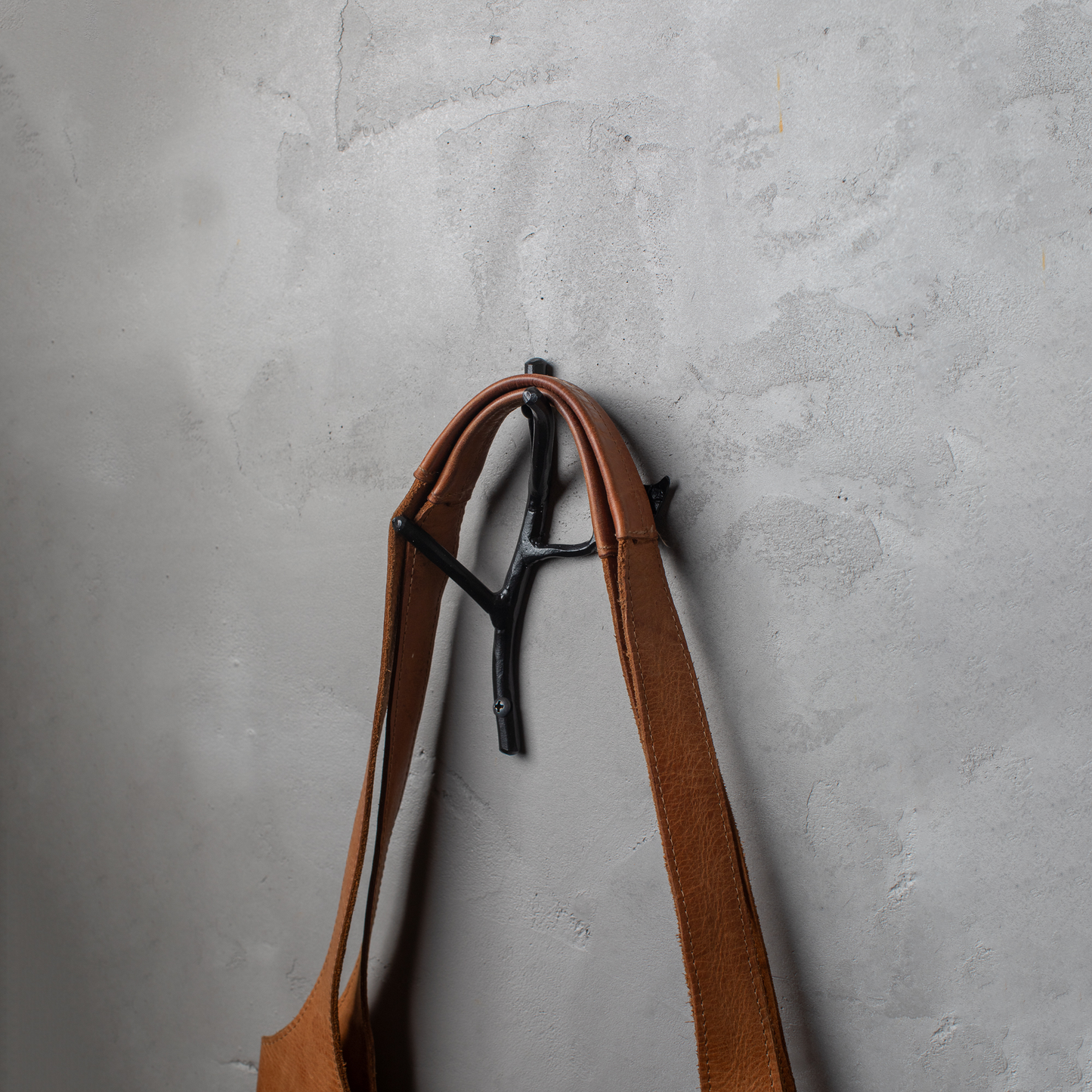 An Urban Forge Sylamore hand-forged iron double hook in a black finish. The hook is mounted on a gray wall and is holding a leather bag.