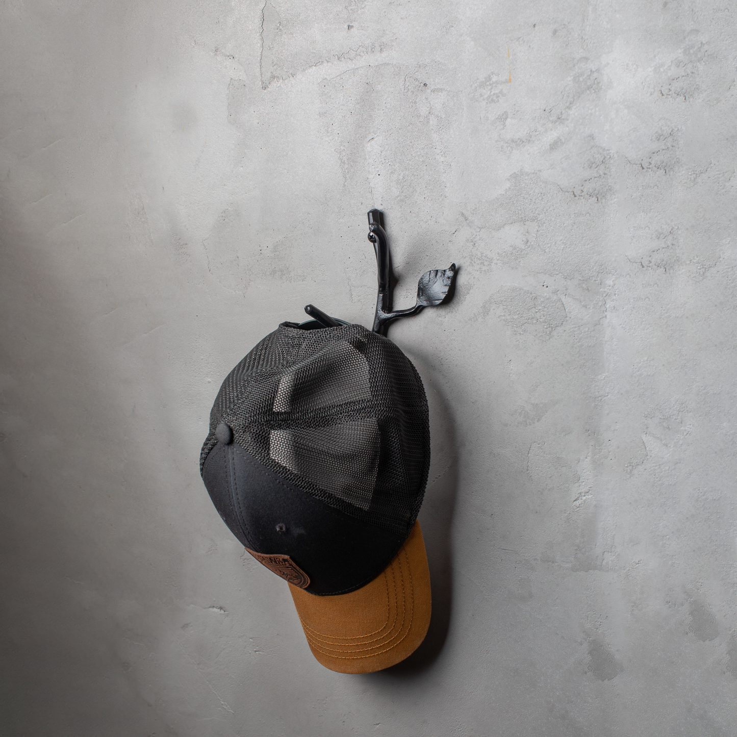 An Urban Forge Sylamore hand-forged iron double hook in a black finish. The hook is mounted on a gray wall and is holding a ball cap.