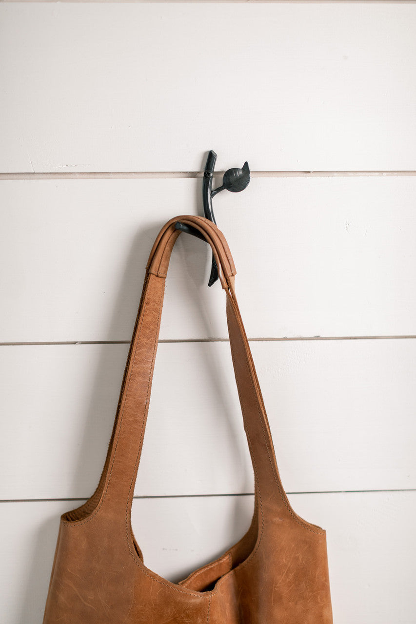 An Urban Forge Sylamore hand-forged iron single hook in a black finish. The hook is mounted on a white shiplap wall and is holding a leather bag.