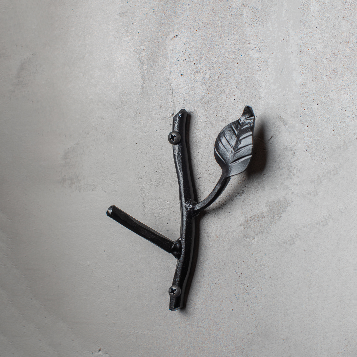 An Urban Forge Sylamore hand-forged iron single hook in a black finish. The hook is mounted on a gray wall.
