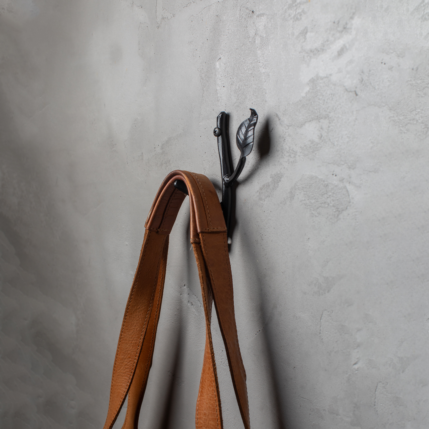 An Urban Forge Sylamore hand-forged iron single hook in a black finish. The hook is mounted on a gray wall and is holding a leather bag.