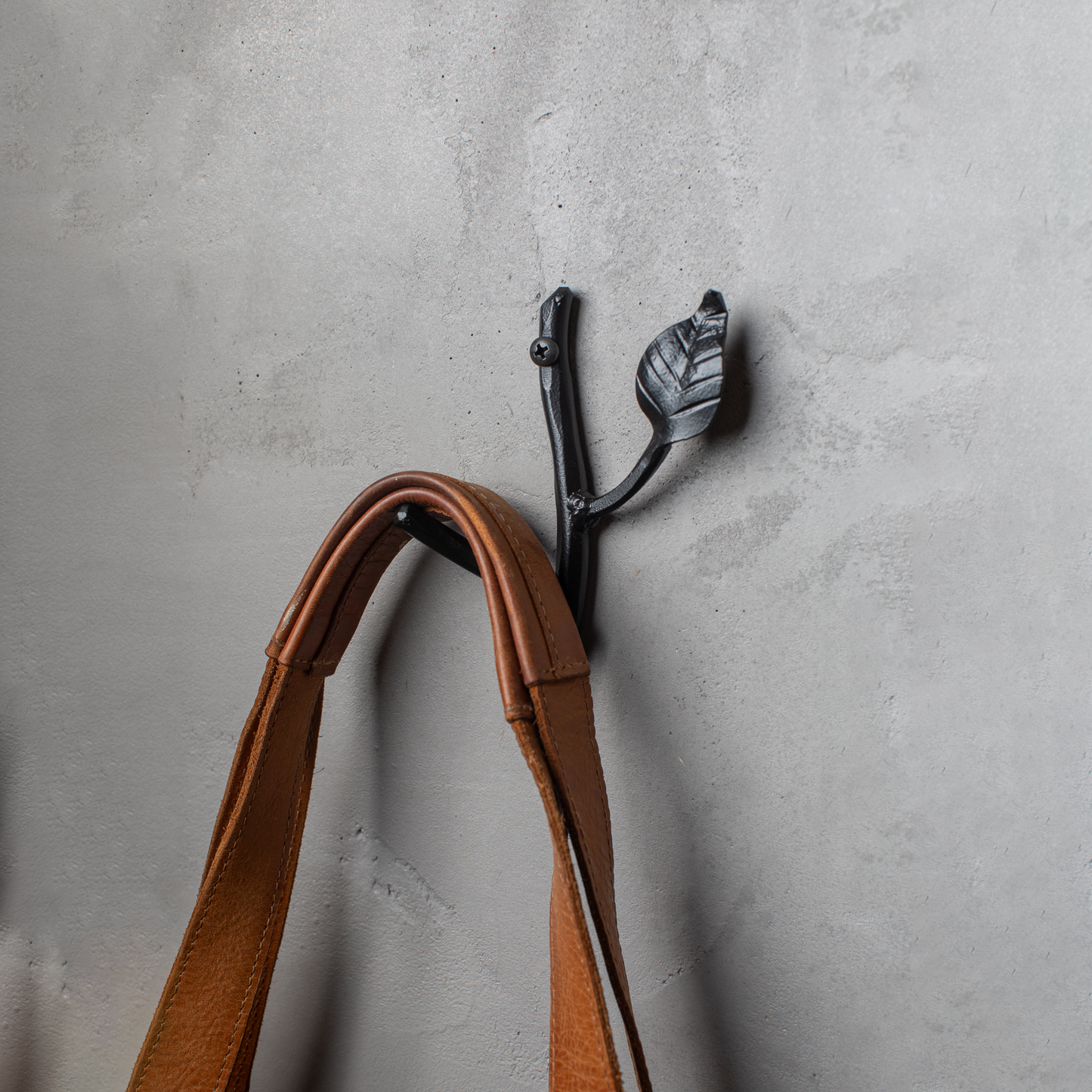 An Urban Forge Sylamore hand-forged iron single hook in a black finish. The hook is mounted on a gray wall and is holding a leather bag.
