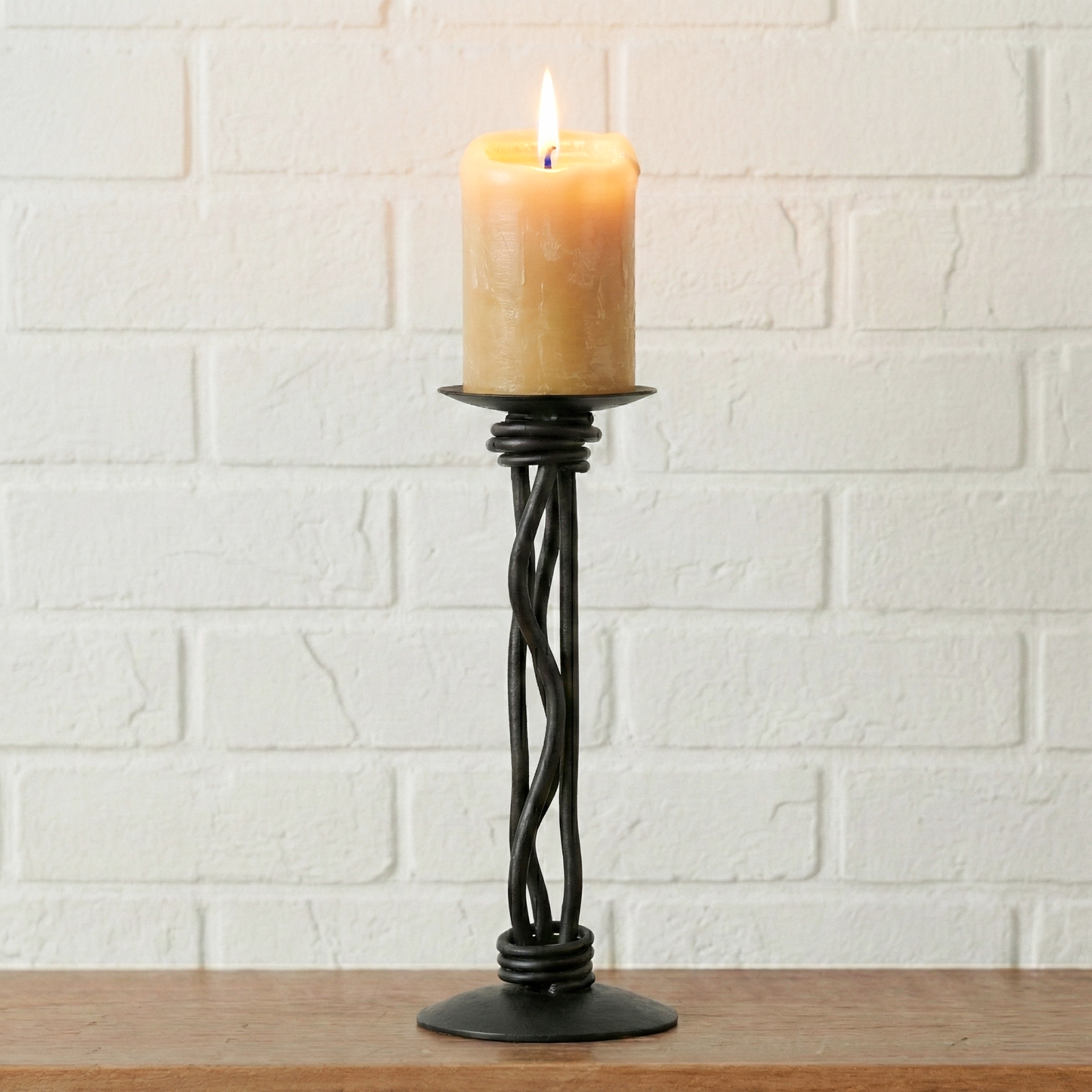 A hand-forged iron candle holder sitting on a fireplace mantle. It is holding a lit candle.