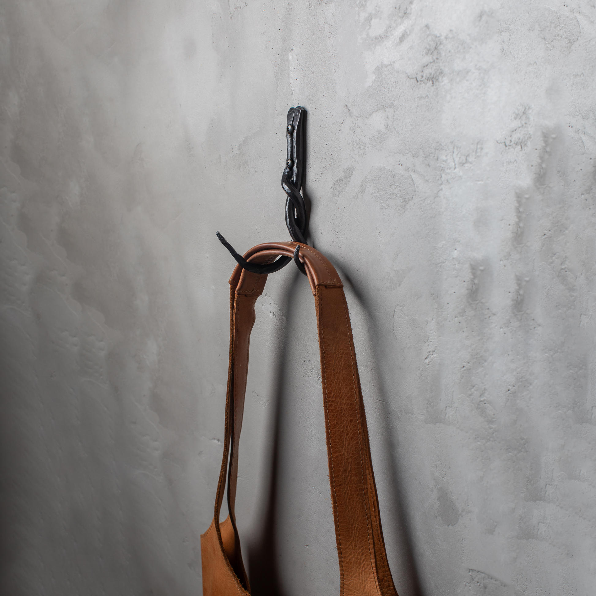An Urban Forge Black River hand-forged iron double hook in a black finish. The hook is mounted on a gray wall and is holding a leather bag.