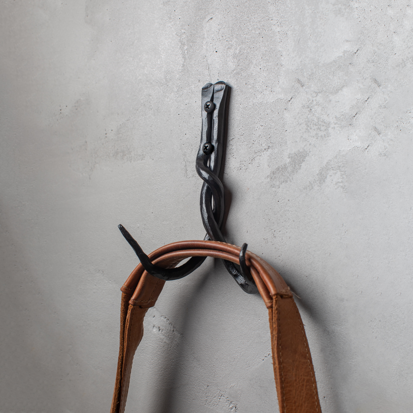 An Urban Forge Black River hand-forged iron double hook in a black finish. The hook is mounted on a gray wall and is holding a leather bag.