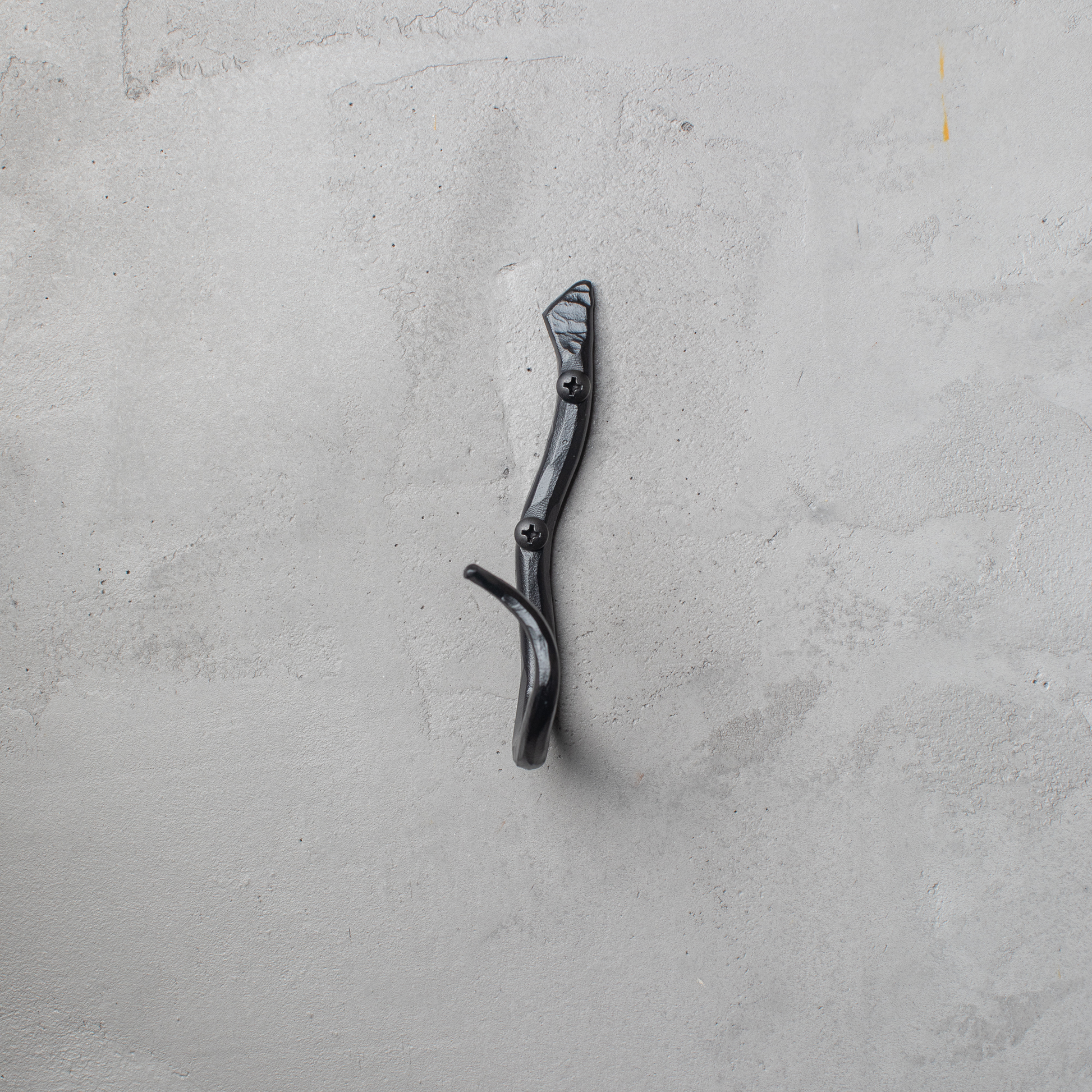An Urban Forge Black River hand-forged iron single hook in a black finish. The hook is mounted on a gray wall.