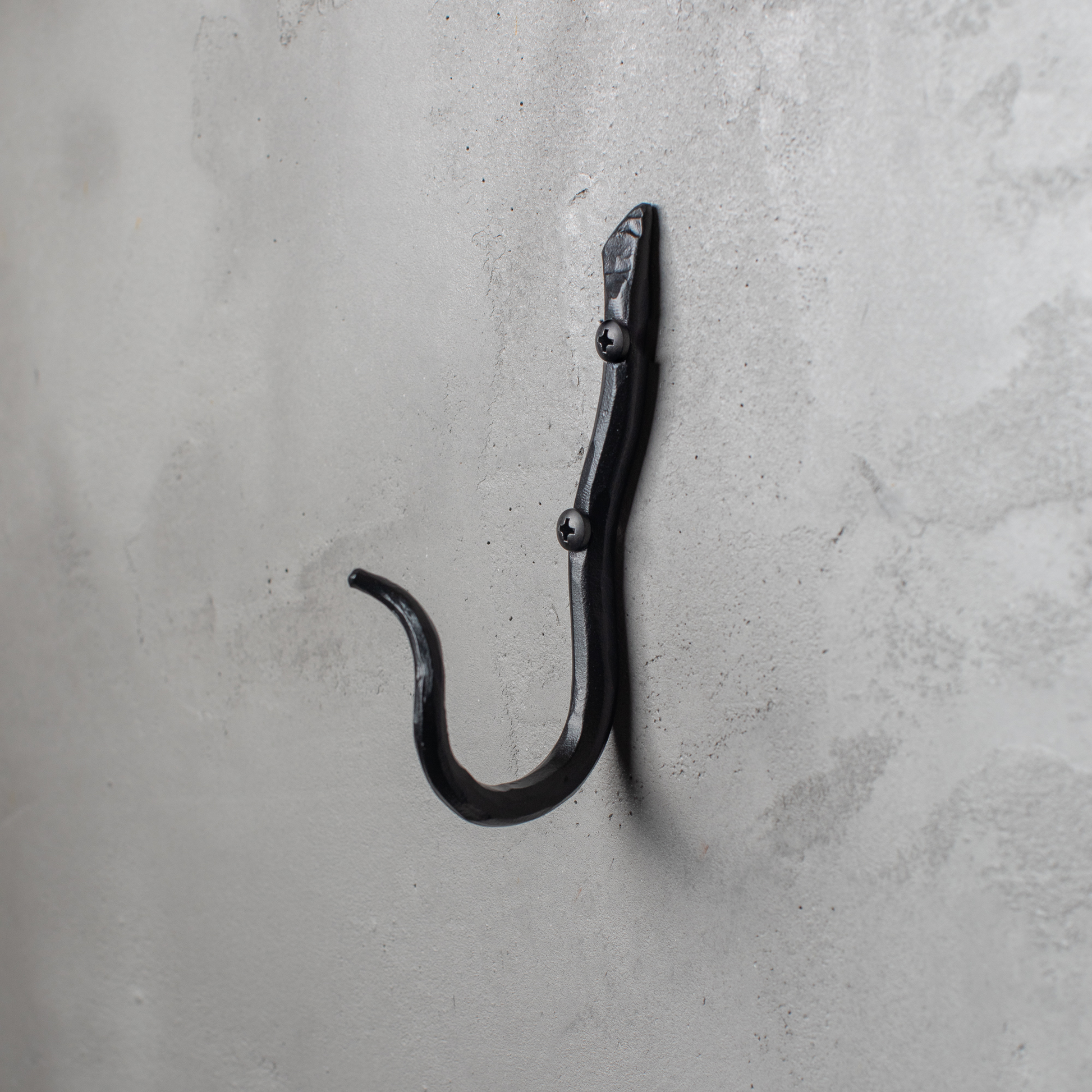 An Urban Forge Black River hand-forged iron single hook in a black finish. The hook is mounted on a gray wall.