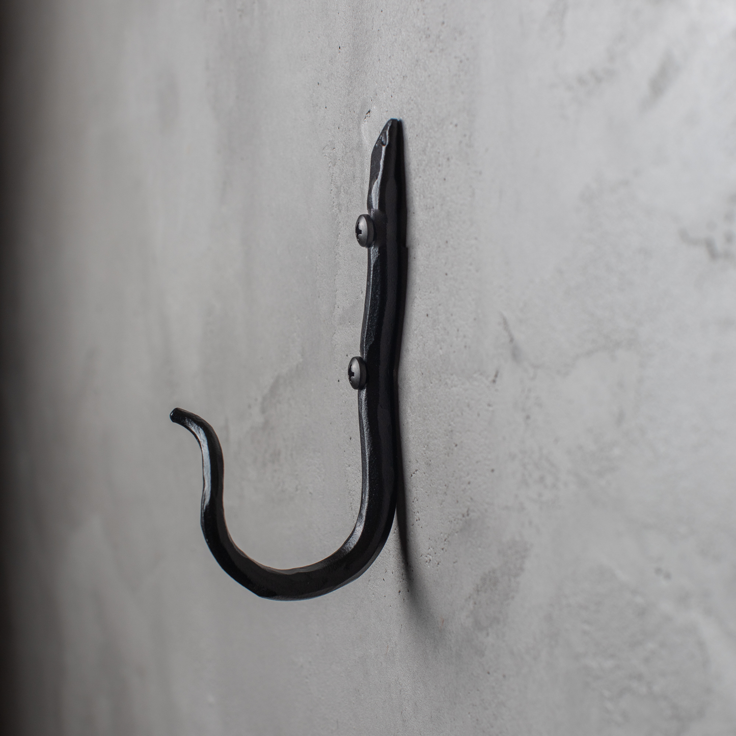An Urban Forge Black River hand-forged iron single hook in a black finish. The hook is mounted on a gray wall.