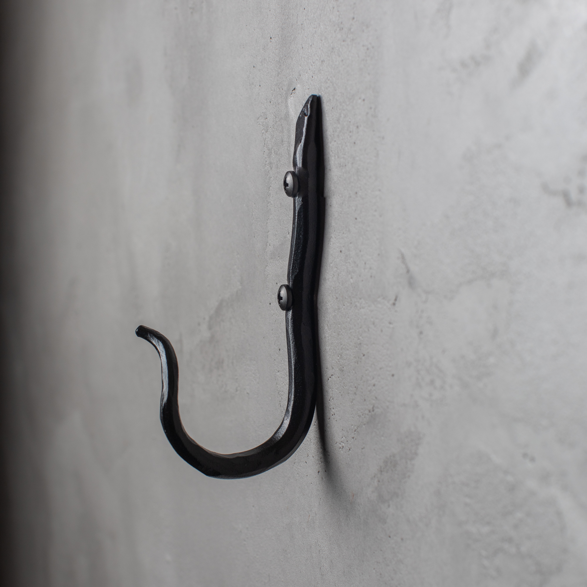 An Urban Forge Black River hand-forged iron single hook in a black finish. The hook is mounted on a gray wall.