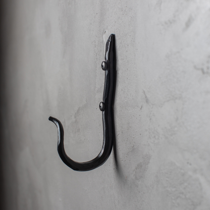 An Urban Forge Black River hand-forged iron single hook in a black finish. The hook is mounted on a gray wall.