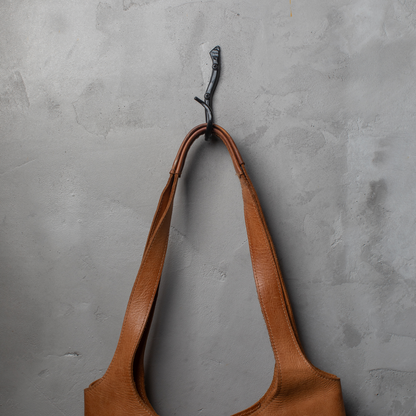 An Urban Forge Black River hand-forged iron single hook in a black finish. The hook is mounted on a gray wall and is holding a leather bag.