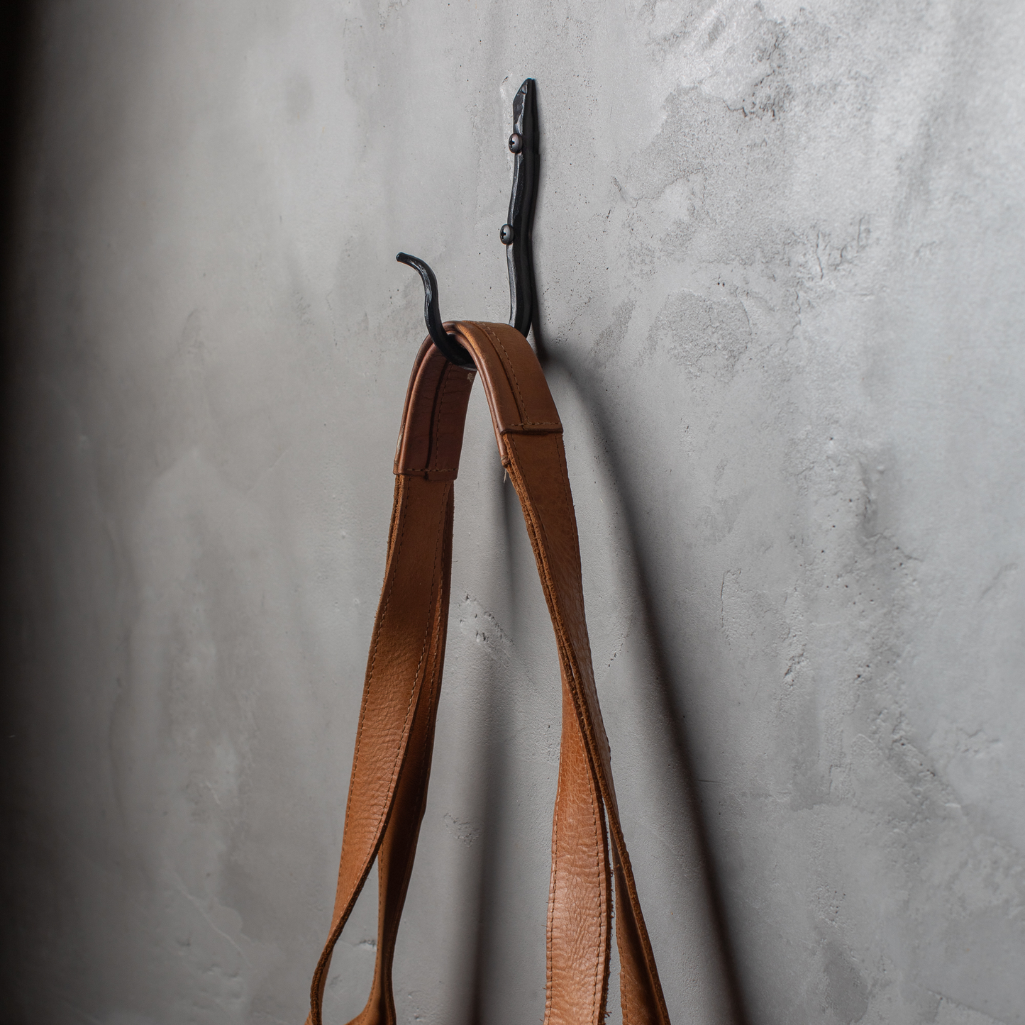An Urban Forge Black River hand-forged iron single hook in a black finish. The hook is mounted on a gray wall and is holding a leather bag.