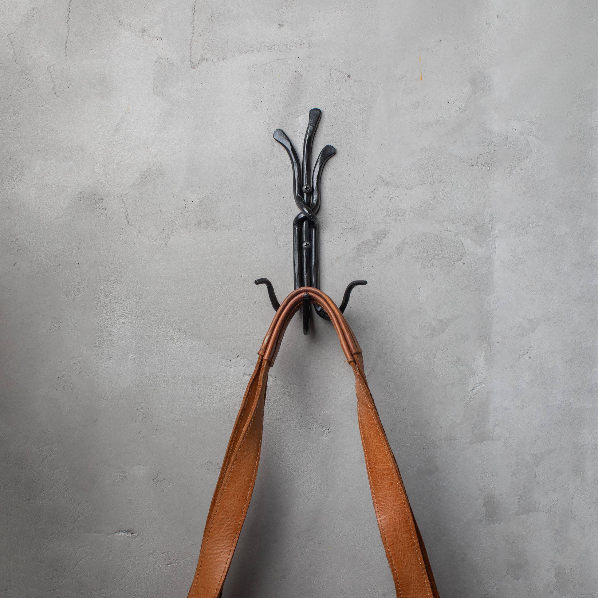 An Urban Forge Black River hand-forged iron triple hook in a black finish. The hook is mounted on a gray wall and is holding a leather bag.