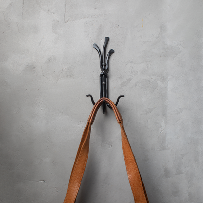 An Urban Forge Black River hand-forged iron triple hook in a black finish. The hook is mounted on a gray wall and is holding a leather bag.