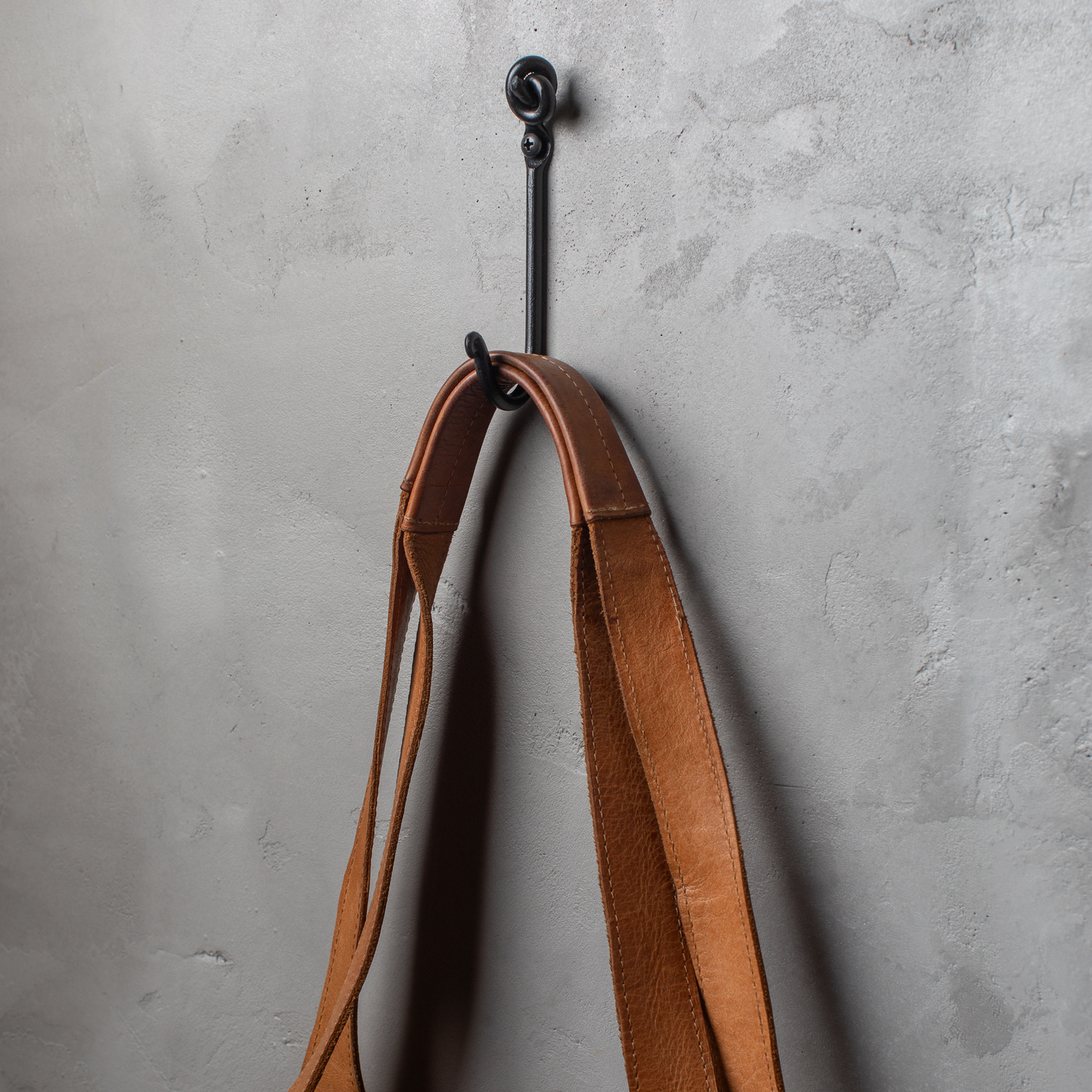 An Urban Forge Knot hand-forged iron single hook in a black finish. The hook is mounted on a gray wall and is holding a leather strap.