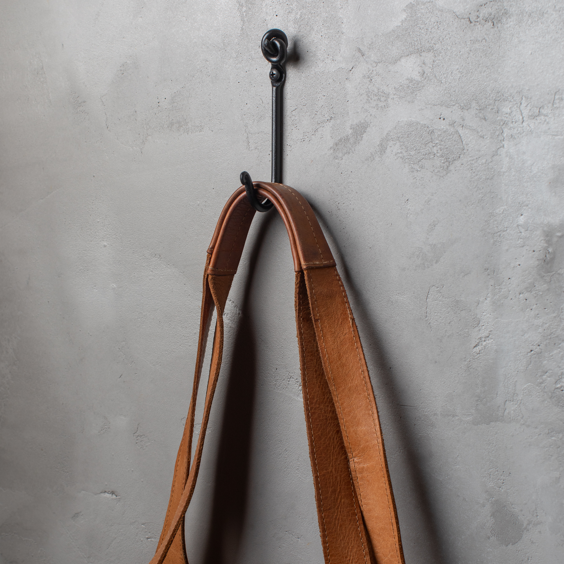 An Urban Forge Knot hand-forged iron single hook in a black finish. The hook is mounted on a gray wall and is holding a leather strap.