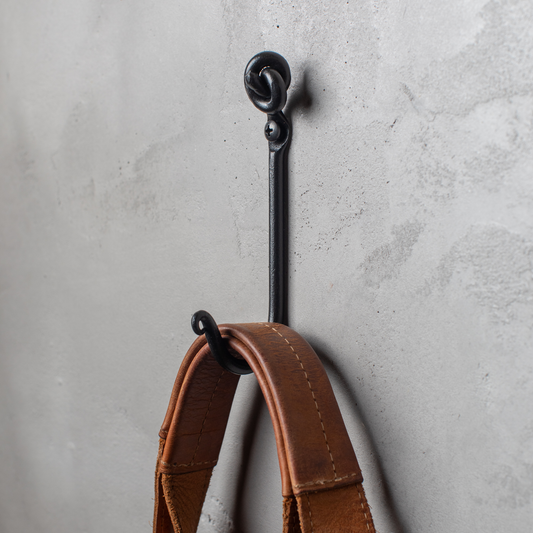 An Urban Forge Knot hand-forged iron single hook in a black finish. The hook is mounted on a gray wall and is holding a leather strap.