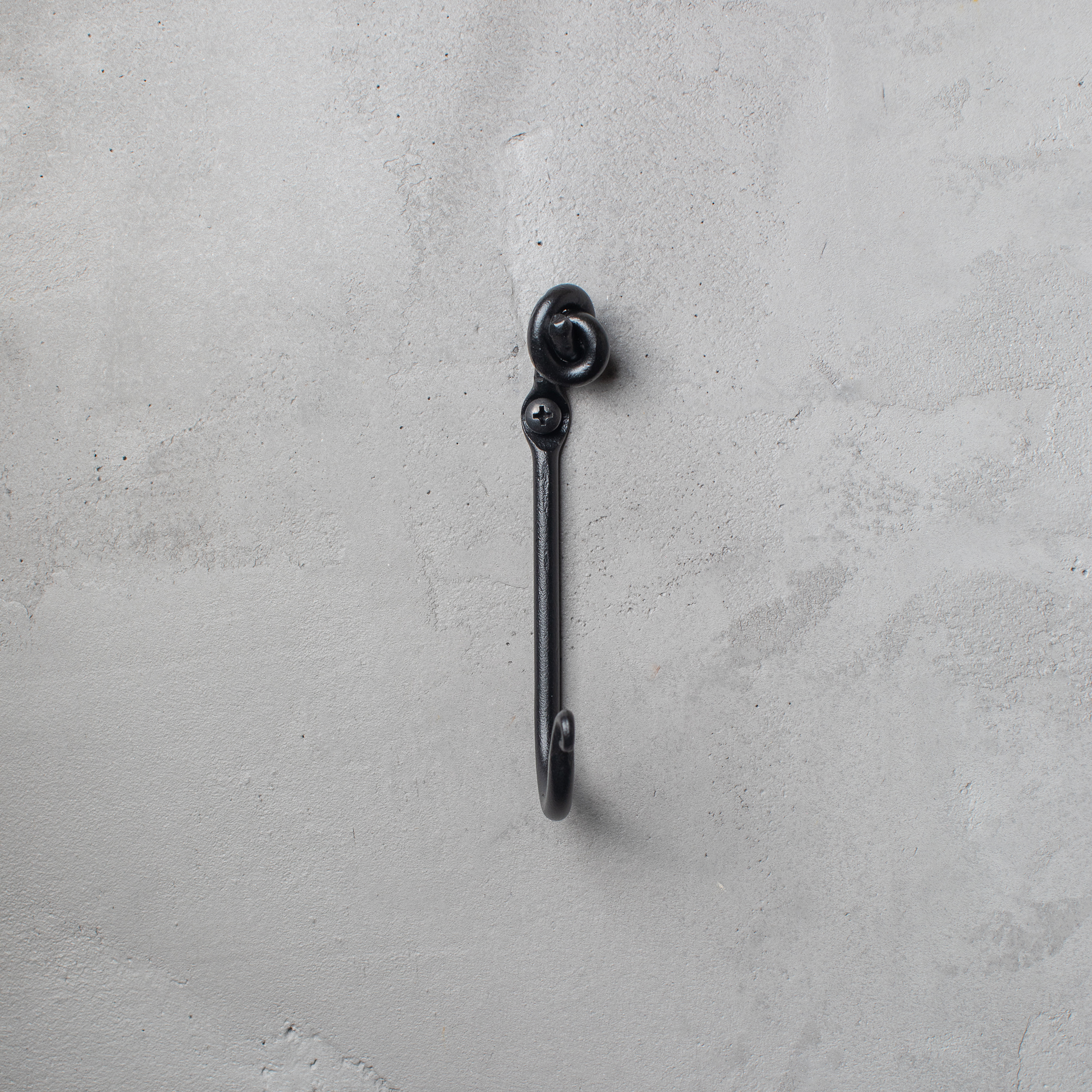 An Urban Forge Knot hand-forged iron single hook in a black finish. The hook is mounted on a gray wall.