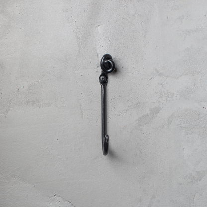 An Urban Forge Knot hand-forged iron single hook in a black finish. The hook is mounted on a gray wall.