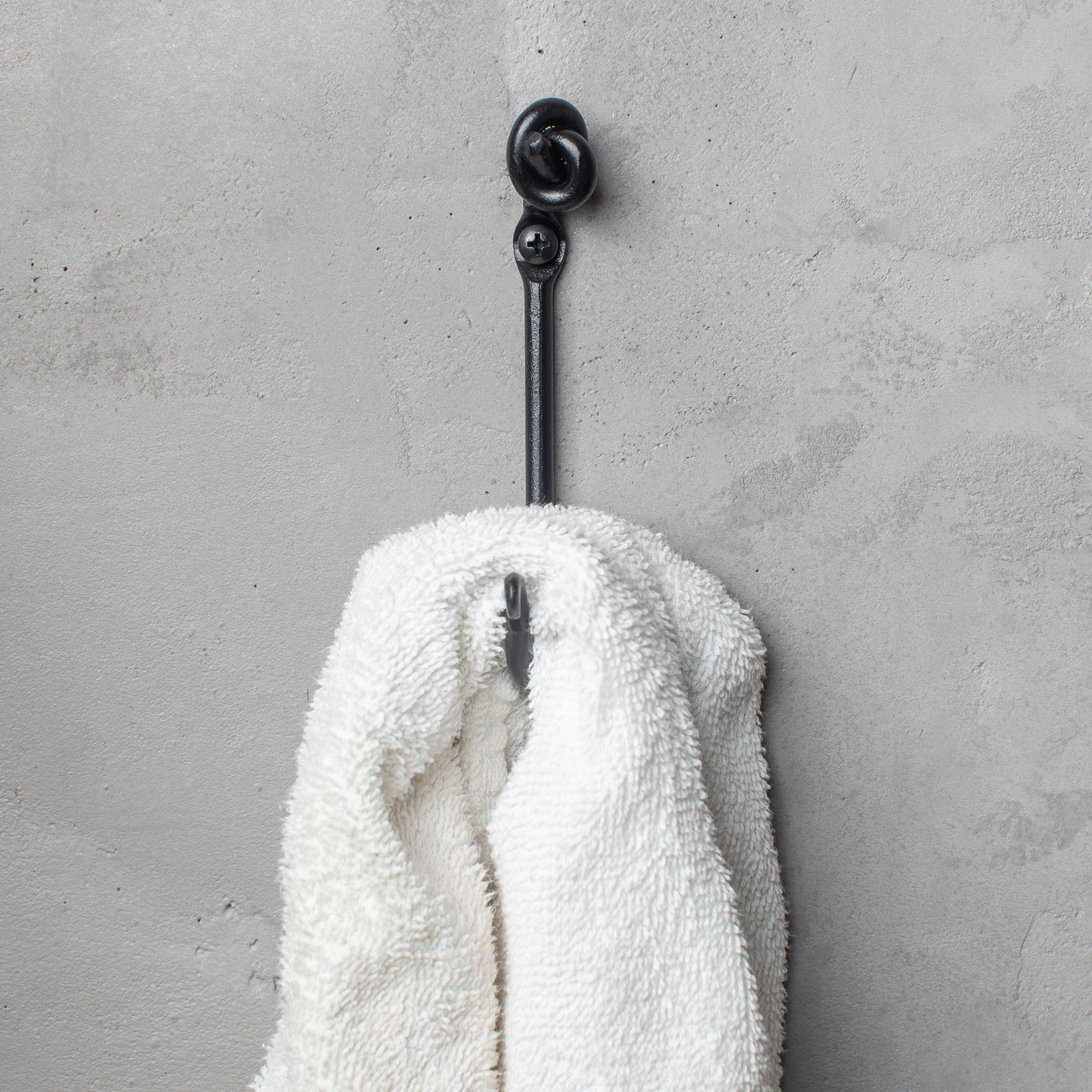 An Urban Forge Knot hand-forged iron single hook in a black finish. The hook is mounted on a gray wall and is holding a white towel.