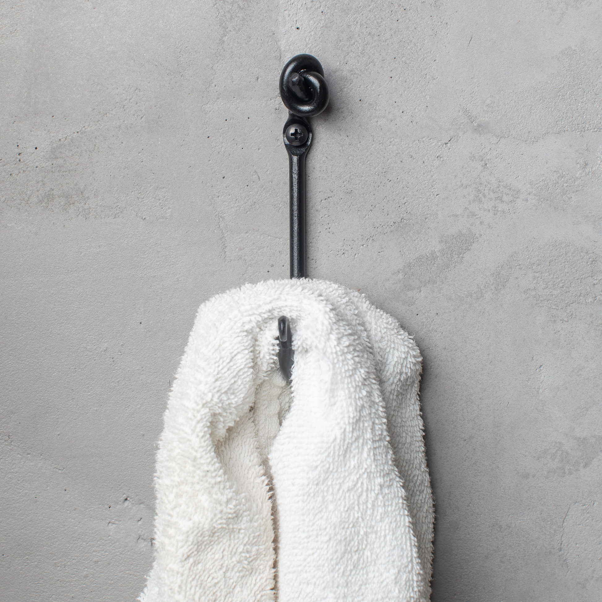 An Urban Forge Knot hand-forged iron single hook in a black finish. The hook is mounted on a gray wall and is holding a white towel.